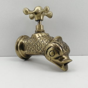 Può includere: Un rubinetto in ottone con un beccuccio a forma di pesce e una maniglia a forma di croce. Il rubinetto presenta un motivo a scaglie dettagliato ed è montato su una base rotonda. L'apparecchio è di un caldo color oro antico.