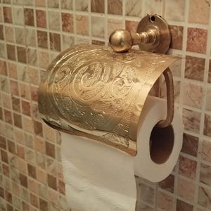 May include: A gold-colored toilet paper holder with an ornate, embossed design. The holder is mounted on a tiled wall with a mosaic pattern of brown, beige, and white squares. A roll of white toilet paper is in place.