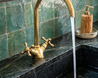 May include: A polished brass kitchen faucet with a high arched spout and two cross-shaped handles. Water flows steadily from the faucet into a dark green marble sink. The faucet features a classic design with a warm metallic finish and visible texture.