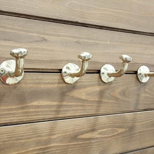 May include: Four brass-colored wall hooks mounted on a horizontal wooden plank background. Each hook has a curved arm and a decorative, rounded top. The wood has a warm, brown tone.