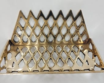 May include: A gold-colored metal fireplace grate with a diamond-shaped pattern. The grate has a pointed top and a flat bottom.