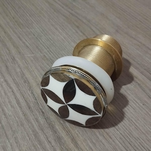 May include: A decorative bottle stopper with a brass base and a white and brown inlaid design. The stopper features a brass neck and a white ring. The inlaid design is a repeating pattern of brown and white shapes.