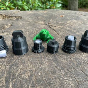 Könnte beinhalten: Sechs schwarze 3D-gedruckte Kappen mit unterschiedlichen Designs, darunter eine Kappe mit einer Schlaufe, eine Kappe mit einem Gewindeloch und eine Kappe mit einer kleinen grünen Froschfigur auf der Oberseite. Die Kappen sind in einer Reihe auf einer Holzoberfläche angeordnet.
