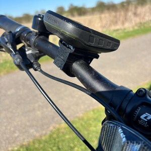 May include: A black Garmin cycling computer mounted on a black bicycle handlebar with a black strap. The computer has a black and green screen with the word "Garmin" printed on it.