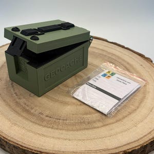 May include: An olive green geocache box with a hinged lid and black details. The box is labelled "GEOCACHE" and is on a wooden surface. A small packet with a log sheet is next to the box.