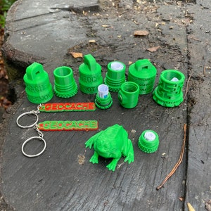 Può includere: Una collezione di contenitori e accessori per geocaching verdi stampati in 3D. L'immagine mostra vari design di contenitori, una statuetta a forma di rana e un portachiavi con la scritta "GEOCACHE". Gli oggetti sono disposti su una superficie di legno stagionato.