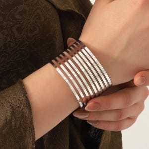 Op de afbeelding: Een zilverkleurige manchetarmband met een verticaal staafontwerp. De armband wordt om een pols gedragen en de persoon draagt een donkergroene blouse. De armband is gemaakt van meerdere parallelle staven.