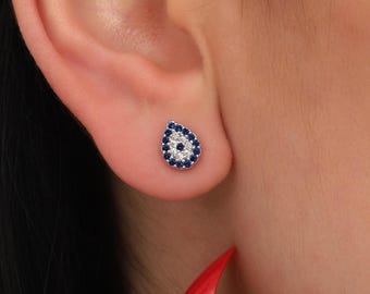 May include: A pair of silver earrings with a teardrop shape and a blue evil eye design. The earrings are set with small white and blue gemstones.