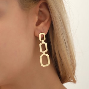 May include: Gold-coloured geometric drop earrings. The earrings feature a series of connected shapes, including rectangles and octagons. The earrings are designed to dangle and catch the light, adding a touch of elegance. A statement piece of jewellery.