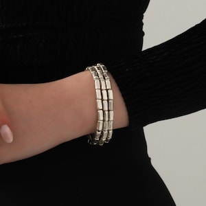 May include: A triple-strand silver-toned bracelet featuring small, rectangular, rounded metal beads strung together in three parallel rows. The bracelet has a polished metallic finish and a uniform design, creating a layered appearance on the wrist.