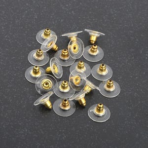 May include: A collection of clear, round earring backs with gold-colored posts. The earring backs are made of a soft, transparent material and are designed to secure earrings. The image shows multiple earring backs scattered on a dark surface.
