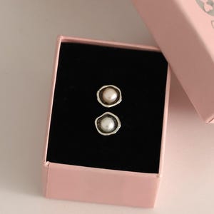 May include: A pair of pearl stud earrings in a pink jewelry box with a black interior. The earrings have a silver-colored bezel setting around each pearl. The box is open, revealing the earrings.