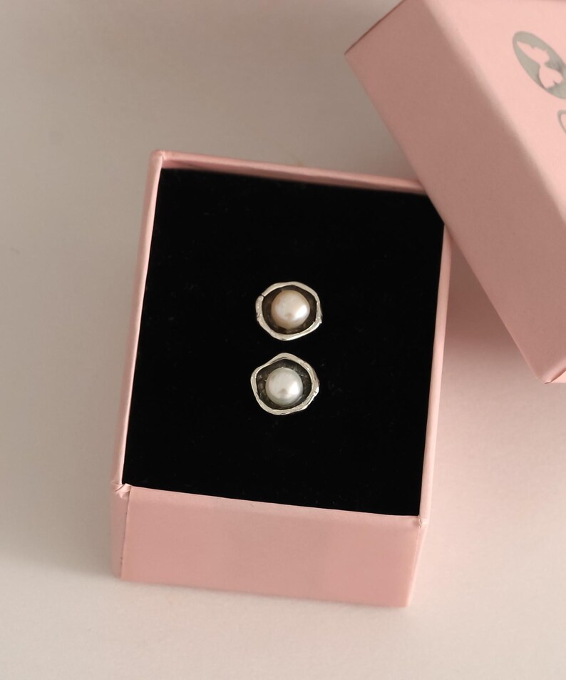 May include: A pair of pearl stud earrings in a pink jewelry box with a black interior. The earrings have a silver-colored bezel setting around each pearl. The box is open, revealing the earrings.