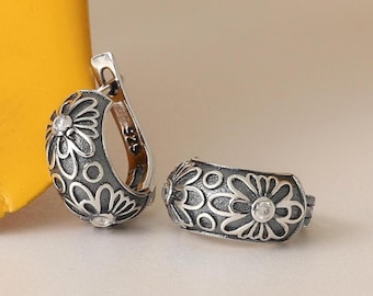 May include: A pair of silver hoop earrings featuring an oxidized finish with raised floral patterns and small clear stones. The interior of the earring post is clearly stamped with the 925 hallmark, indicating sterling silver composition.