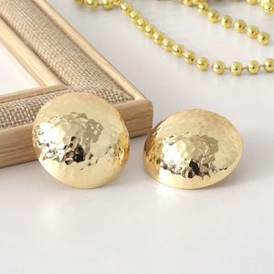 May include: Two gold-colored, hammered-metal earrings with a domed shape. The earrings are next to a gold beaded necklace and a wooden picture frame with a burlap-like texture.