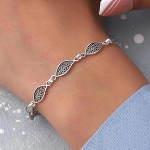 May include: A silver-toned bracelet featuring a series of interconnected, leaf-shaped links. Each link contains an intricate, openwork filigree pattern. The bracelet is secured with small metal jump rings connecting the individual decorative elements.