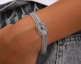 May include: A silver-toned bracelet featuring a woven chain design that converges into a central knot detail. The bracelet is constructed from multiple strands of fine, textured metal links that form a continuous, flexible band around the wrist.