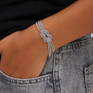 Puede incluir: Un brazalete de plata con un dise&ntilde;o anudado. El brazalete est&aacute; hecho de m&uacute;ltiples hebras de cadena de plata.