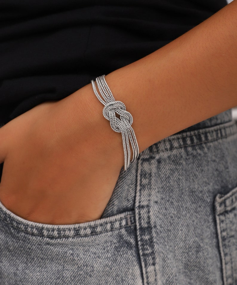 Puede incluir: Un brazalete de plata con un dise&ntilde;o anudado. El brazalete est&aacute; hecho de m&uacute;ltiples hebras de cadena de plata.