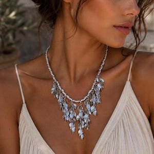 May include: A silver-toned necklace featuring a chain of small round beads and a central cluster of dangling leaf-shaped charms. The metallic leaves are arranged in multiple layers, creating a cascading effect that rests against the collarbone area.
