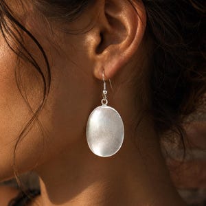 May include: A silver-toned oval drop earring featuring a smooth, slightly concave surface with a subtle metallic sheen. The earring is suspended from a simple silver-colored metal hook wire that passes through the earlobe for secure attachment.