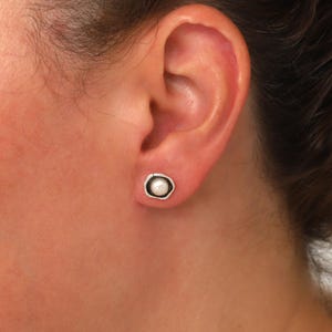 May include: A close-up of a pearl stud earring. The earring features a small, round, white pearl set in a silver-colored, irregular bezel. The earring is worn on an ear.