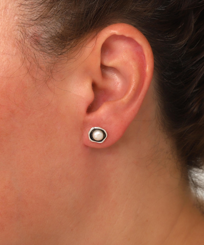 May include: A close-up of a pearl stud earring. The earring features a small, round, white pearl set in a silver-colored, irregular bezel. The earring is worn on an ear.