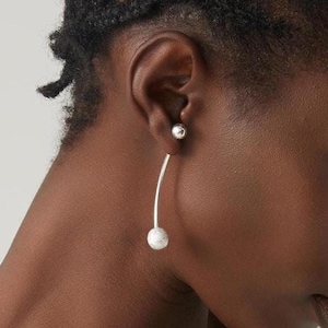 May include: A silver-toned earring featuring a small, smooth spherical stud at the front of the earlobe, connected to a curved silver bar that extends downward, ending in a larger, textured spherical bead that sits below the earlobe.