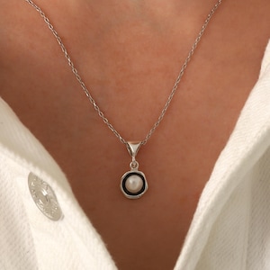 May include: A silver necklace with a small pendant featuring a white pearl surrounded by a black border. The pendant hangs from a delicate chain. The necklace is displayed against a white background.