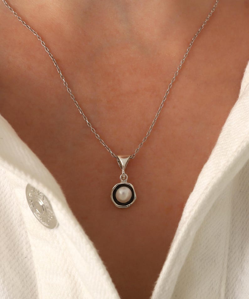 May include: A silver necklace with a small pendant featuring a white pearl surrounded by a black border. The pendant hangs from a delicate chain. The necklace is displayed against a white background.