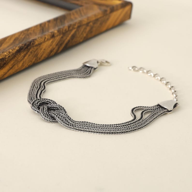 Puede incluir: Un brazalete de plata con un dise&ntilde;o anudado y m&uacute;ltiples eslabones de cadena. El brazalete tiene un cierre de broche.