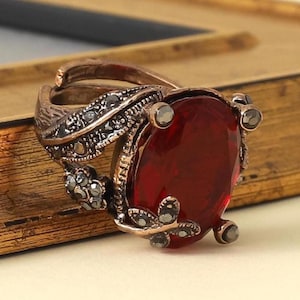 May include: A copper-toned ring featuring a large, oval-shaped, deep red faceted stone at the center. The band and setting are decorated with small, dark, metallic accents arranged in leaf-like patterns surrounding the central red stone.