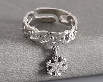 May include: A silver-toned metal ring featuring a chain-link band design with an open back. A small, dangling snowflake-shaped charm is attached to the centre of the band, encrusted with tiny, clear, sparkling stones that catch the light.