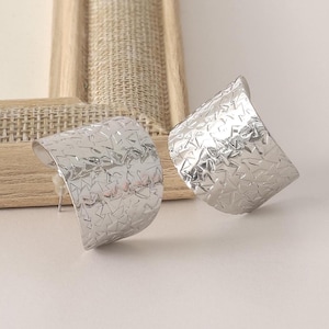 Op de afbeelding: Een paar zilverkleurige, getextureerde oorbellen. Elke oorbel heeft een gebogen, rechthoekige vorm met een geometrisch patroon. De oorbellen worden gepresenteerd op een lichtgekleurd oppervlak.