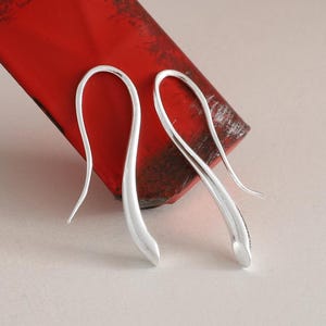 Op de afbeelding: Een paar oorbellen van gepolijst zilver met een glad, langwerpig en gebogen haakontwerp. De oorbellen hebben een strakke, reflecterende metalen afwerking en een doorlopende, vloeiende vorm die onderaan in een punt uitloopt.