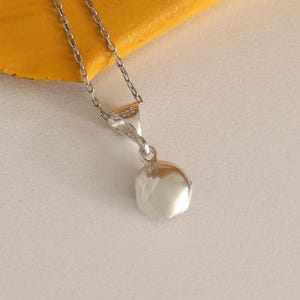 May include: A silver necklace with a small, round pendant. The pendant is a shiny, reflective silver color. The chain is delicate and also silver. The necklace is displayed on a yellow surface.