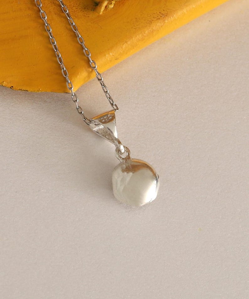May include: A silver necklace with a small, round pendant. The pendant is a shiny, reflective silver color. The chain is delicate and also silver. The necklace is displayed on a yellow surface.