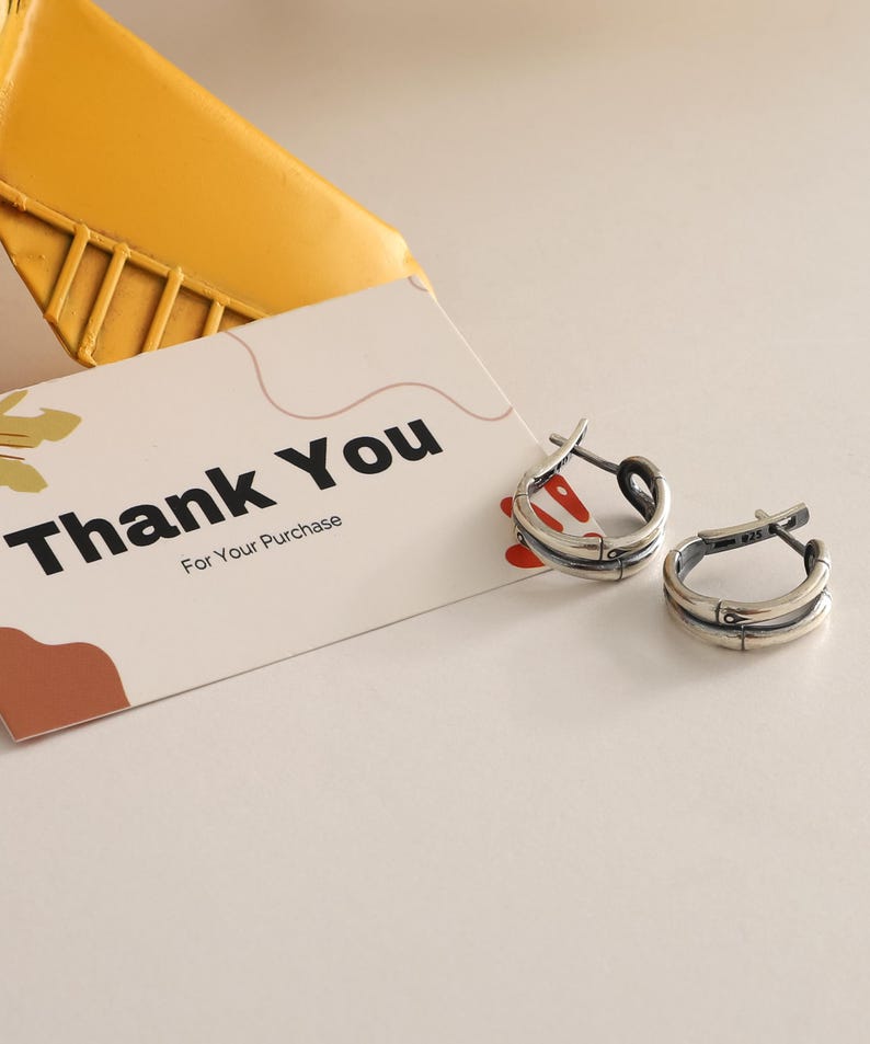 Op de afbeelding: Een paar zilveren oorringen met een textuurontwerp. De oorbellen liggen naast een kaart met de tekst "Thank You" in zwarte tekst. De kaart ligt op een wit oppervlak.