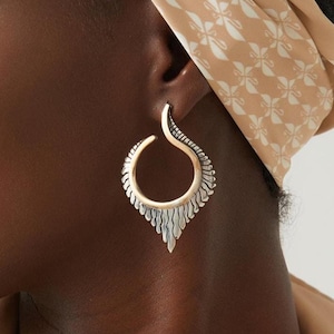 May include: A silver-toned hoop earring featuring a curved, tapered top section and a textured, fan-like lower edge that extends into a pointed base. The metal surface displays a polished finish with darkened recessed details along the outer rim.