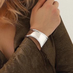 Könnte beinhalten: Ein breites, poliertes silbernes Armband. Das Armband hat ein geschwungenes Design und wird am Handgelenk getragen. Das Metall reflektiert das Licht und erzeugt ein helles, glänzendes Aussehen. Der Schmuck ist ein schlichtes, elegantes Accessoire.