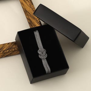 Puede incluir: Un brazalete de cadena de plata con un dise&ntilde;o anudado, presentado en una caja de terciopelo negro.