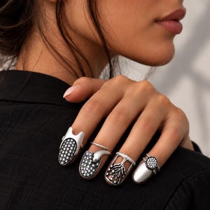 May include: A set of four silver-toned nail rings worn on the fingers. Two rings feature a pavé-style surface with small clear stones, one ring displays a tree-like design with star accents, and one ring has a smooth surface with a circular stone cluster.
