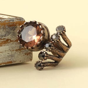 Op de afbeelding: Een bronskleurige ring met een grote, gefacetteerde, ronde bruine edelsteen in een kroonvormige zetting, vergezeld van vier kleinere, donkere, ronde stenen gemonteerd op getextureerde, takachtige metalen pootjes die uit de basis komen.