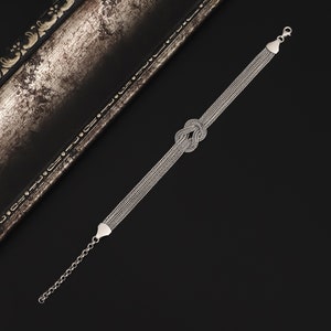 Puede incluir: Un brazalete de plata con un dise&ntilde;o anudado. El brazalete est&aacute; hecho de m&uacute;ltiples hebras de cadena.