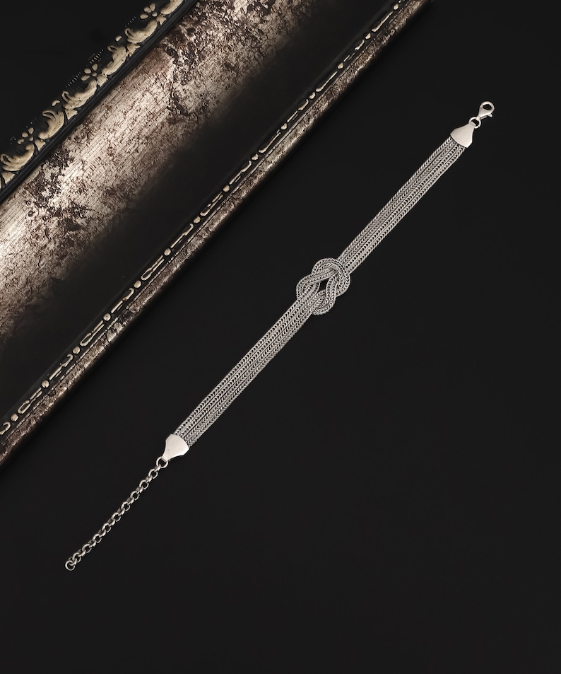 Puede incluir: Un brazalete de plata con un dise&ntilde;o anudado. El brazalete est&aacute; hecho de m&uacute;ltiples hebras de cadena.