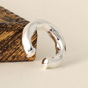 May include: A silver-toned ear cuff with a polished, reflective finish and an irregular, organic, hammered texture. The piece features a C-shaped design with rounded, smooth ends, creating a continuous, flowing form that curves around the ear.
