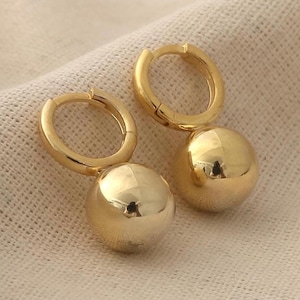 May include: A pair of gold-toned hoop earrings, each featuring a smooth, polished spherical charm suspended from a circular hinged hoop. The metal surface is reflective and uniform in color, showcasing a simple design with no additional embellishments.