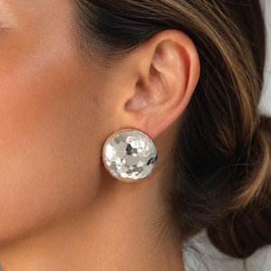 May include: A large, circular stud earring with a polished silver finish and a distinct hammered texture. The surface of the earring features numerous small, concave indentations that catch the light, creating a reflective and multifaceted appearance.