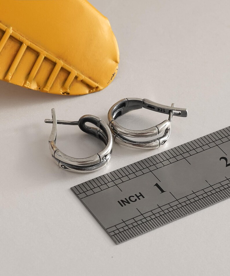 Op de afbeelding: Twee zilveren oorringen met een bamboe-achtig ontwerp worden getoond op een wit oppervlak naast een liniaal. De oorbellen hebben een scharniersluiting. Een geel object bevindt zich op de achtergrond.