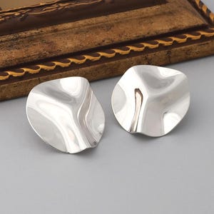 May include: A pair of silver-toned, abstract-shaped earrings. The earrings have a wavy, organic design, reflecting light and creating highlights. The background is a solid gray, with a glimpse of a decorative gold-framed object in the upper portion of the image.
